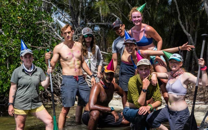 A group of people pose for a photo while standing in shallow water. Some are holding paddles. Some are wearing party hats. 